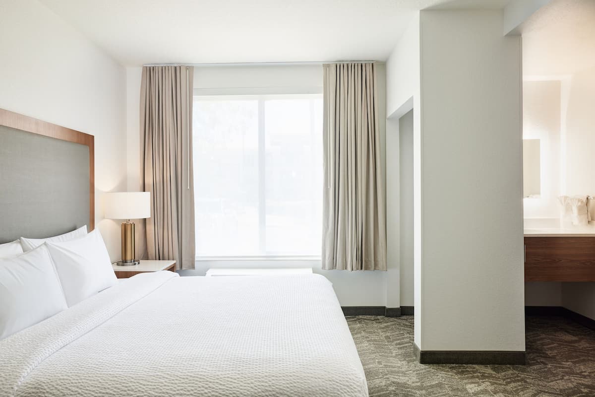 A hotel room with a large bed, a nightstand with a lamp, a window with curtains, and a wooden headboard.