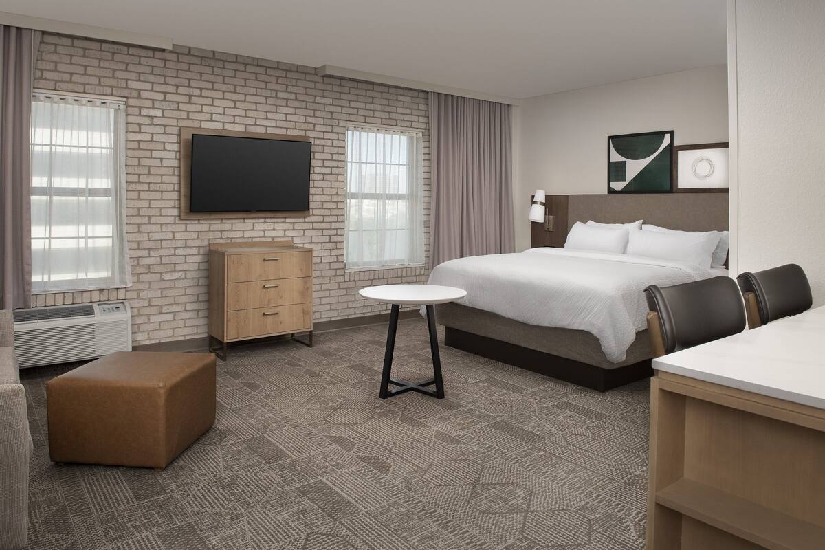 A hotel room with a brick wall, a flat-screen TV, a bed with white bedding, a wooden dresser, and a small round table.