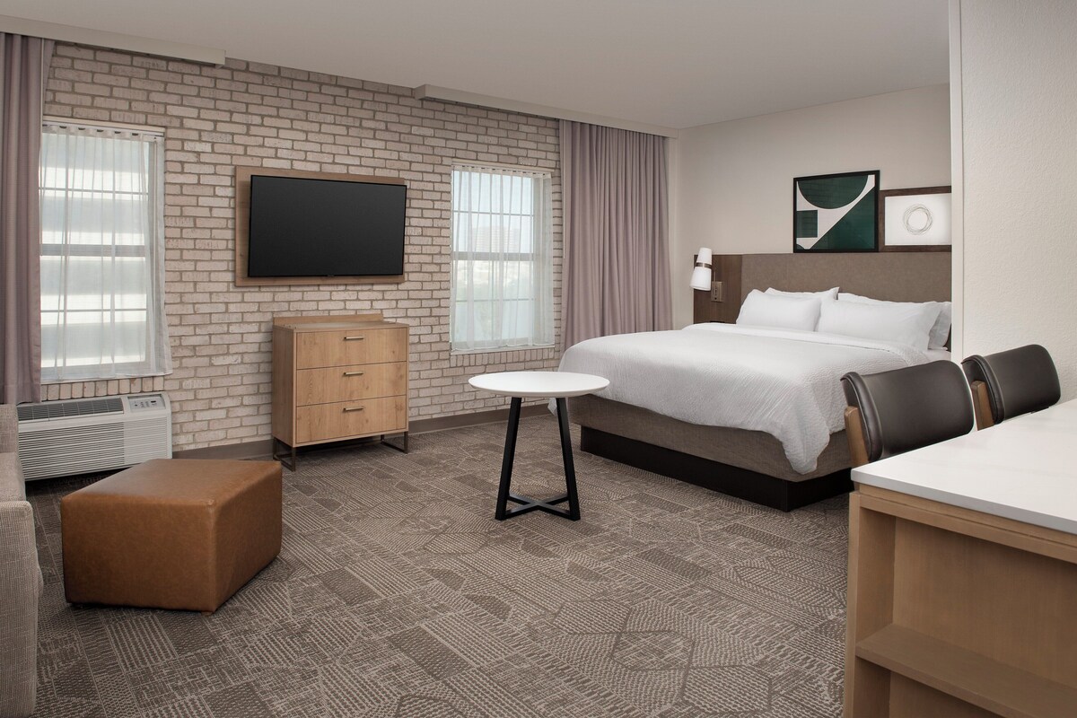 A hotel room with a brick wall, a flat-screen TV, a bed with white bedding, a wooden dresser, and a small round table.