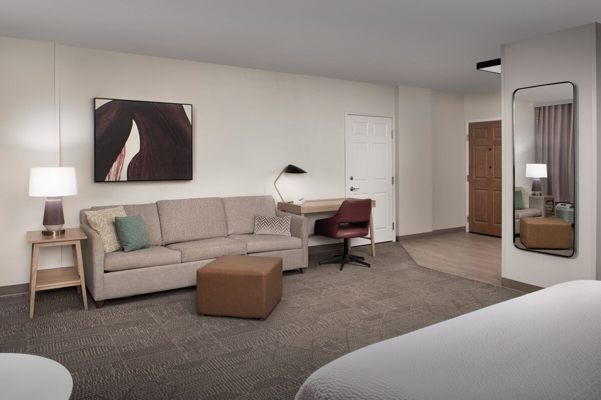 Ein Hotelzimmer mit Sofa, Schreibtisch, Stuhl, Pouf, Lampe, Gemälde und Spiegel.