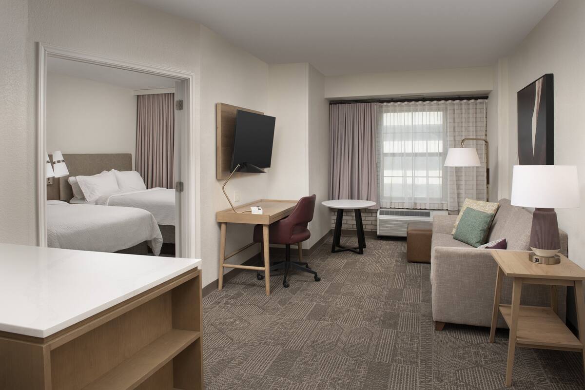 A hotel room with a bed, a desk, a chair, a sofa, a TV mounted on the wall, and a window with curtains.