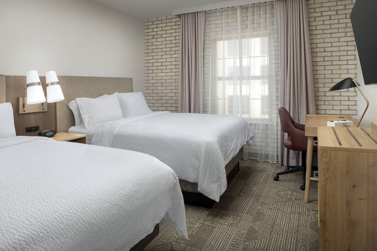 A hotel room with two beds, a desk, a chair, a lamp, and a window with curtains.