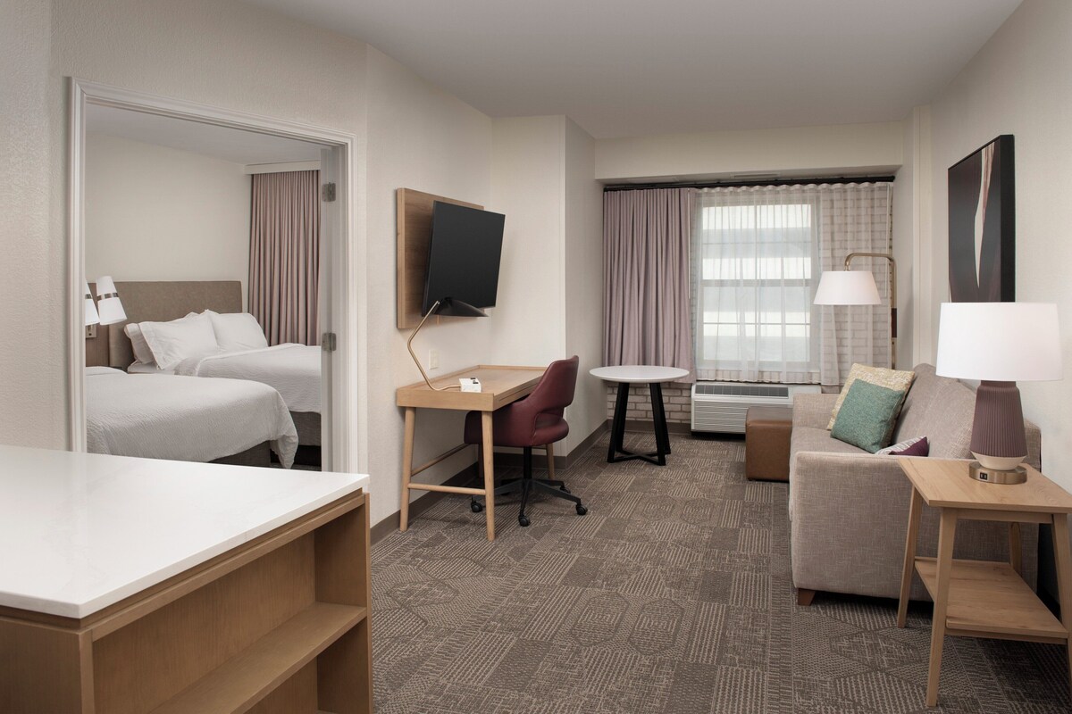 Une chambre d’hôtel comprenant un lit, un bureau, une chaise, un canapé, un téléviseur fixé au mur et une fenêtre avec des rideaux.