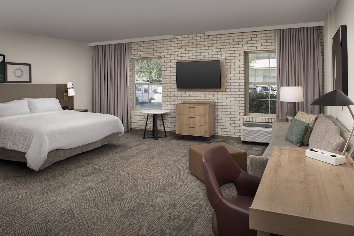 A hotel room with a large bed, a desk with a lamp, a TV mounted on the wall, and a window with curtains.