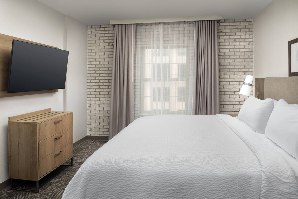 A hotel room with a bed, a TV mounted on the wall, a wooden dresser, and a brick wall.