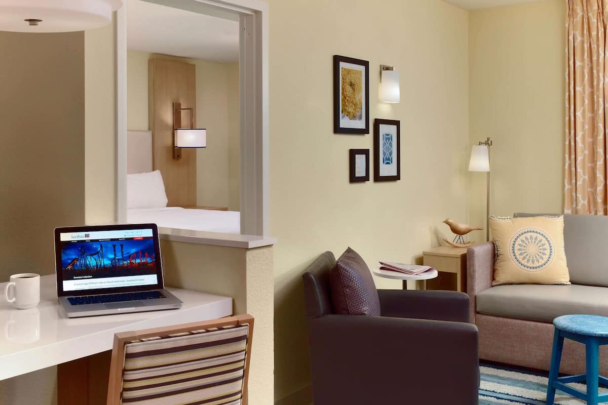 A hotel room with a desk, laptop, chair, sofa, and a small table with a lamp.