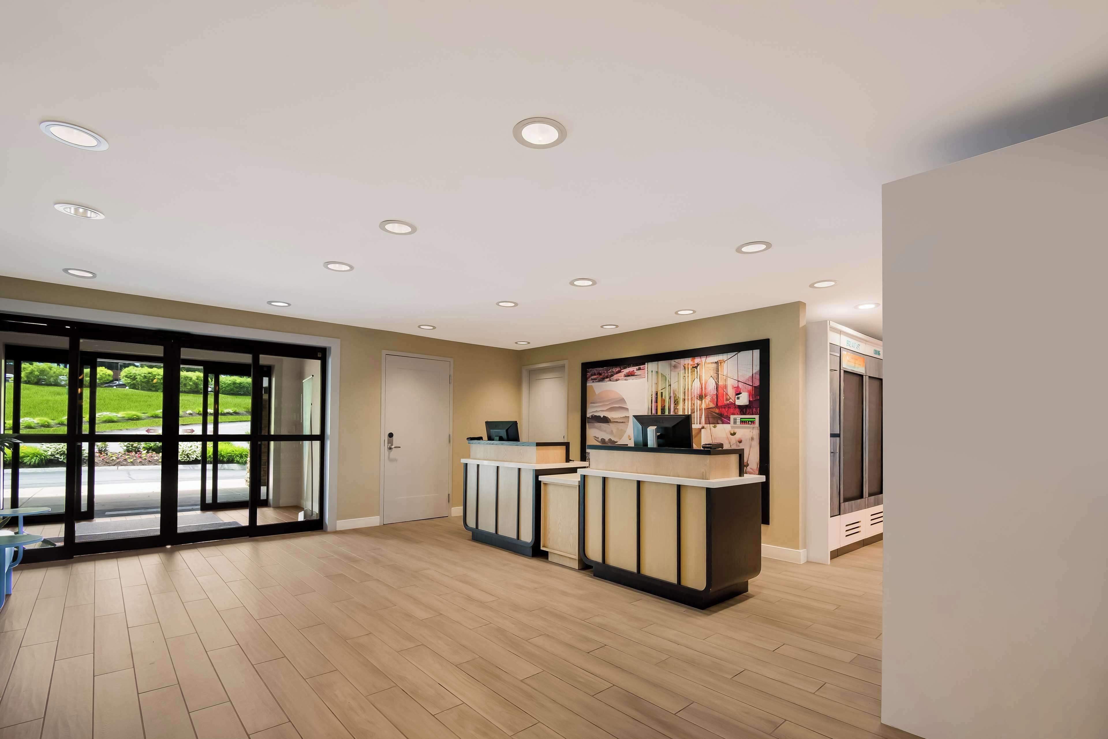 A reception area with a modern design, featuring a reception desk, a large window with a view of greenery, and a ceiling with recessed lighting.
