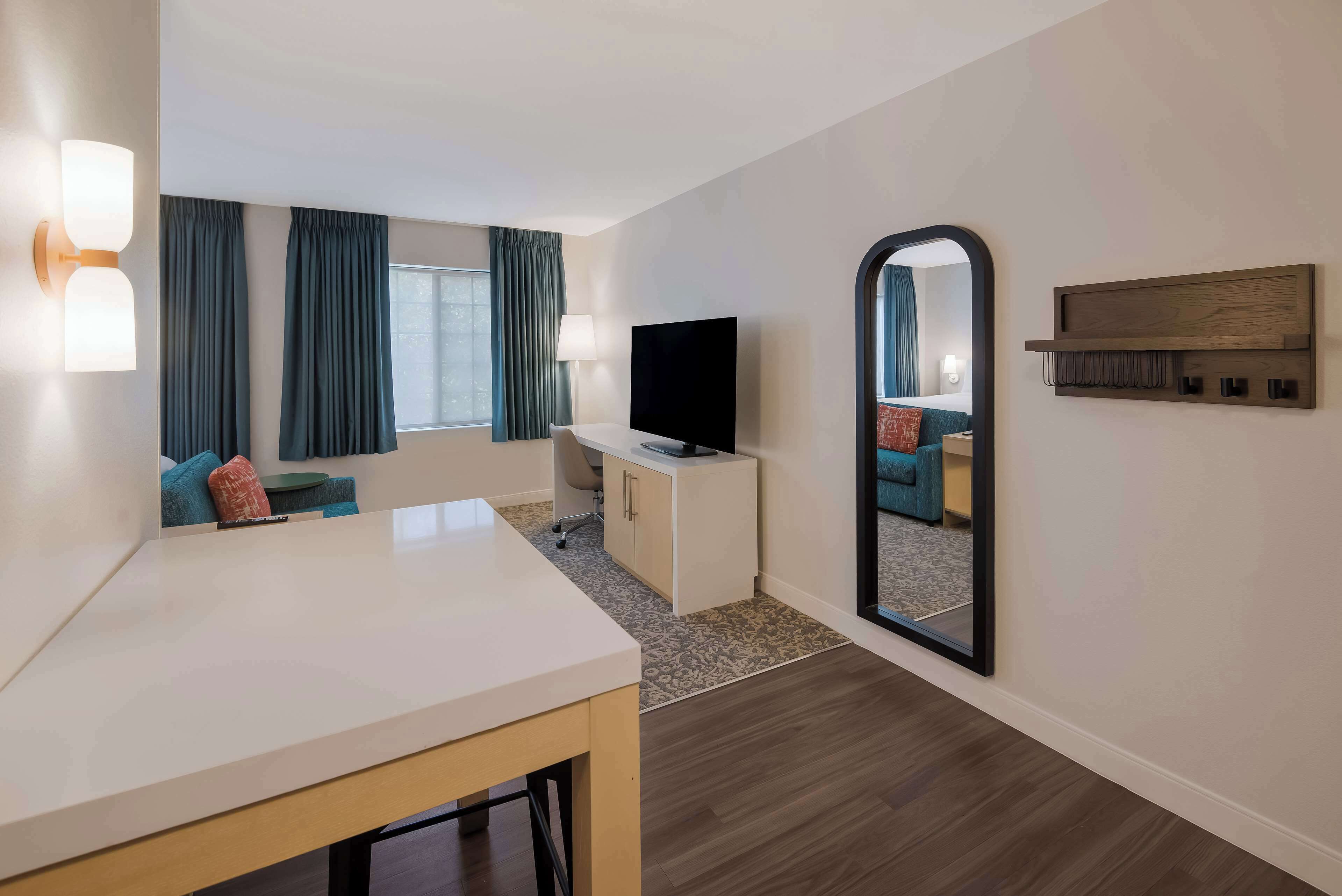 Habitación de hotel con un escritorio blanco, un televisor de pantalla plana, un sofá con cojines azules, un espejo grande y una estantería de madera.
