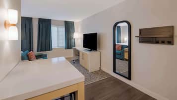 A hotel room with a white desk, a flat-screen TV, a sofa with blue cushions, a large mirror, and a wooden shelf.