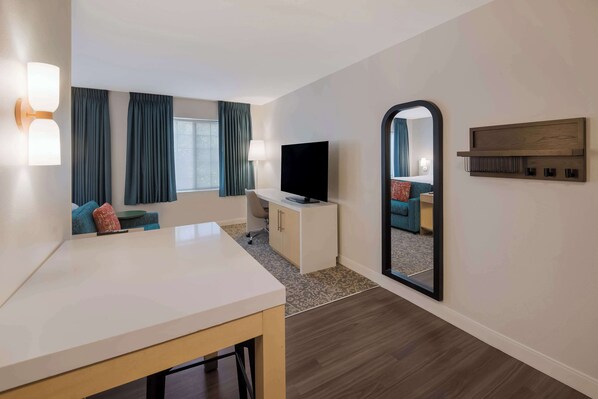 Habitación de hotel con un escritorio blanco, un televisor de pantalla plana, un sofá con cojines azules, un espejo grande y una estantería de madera.