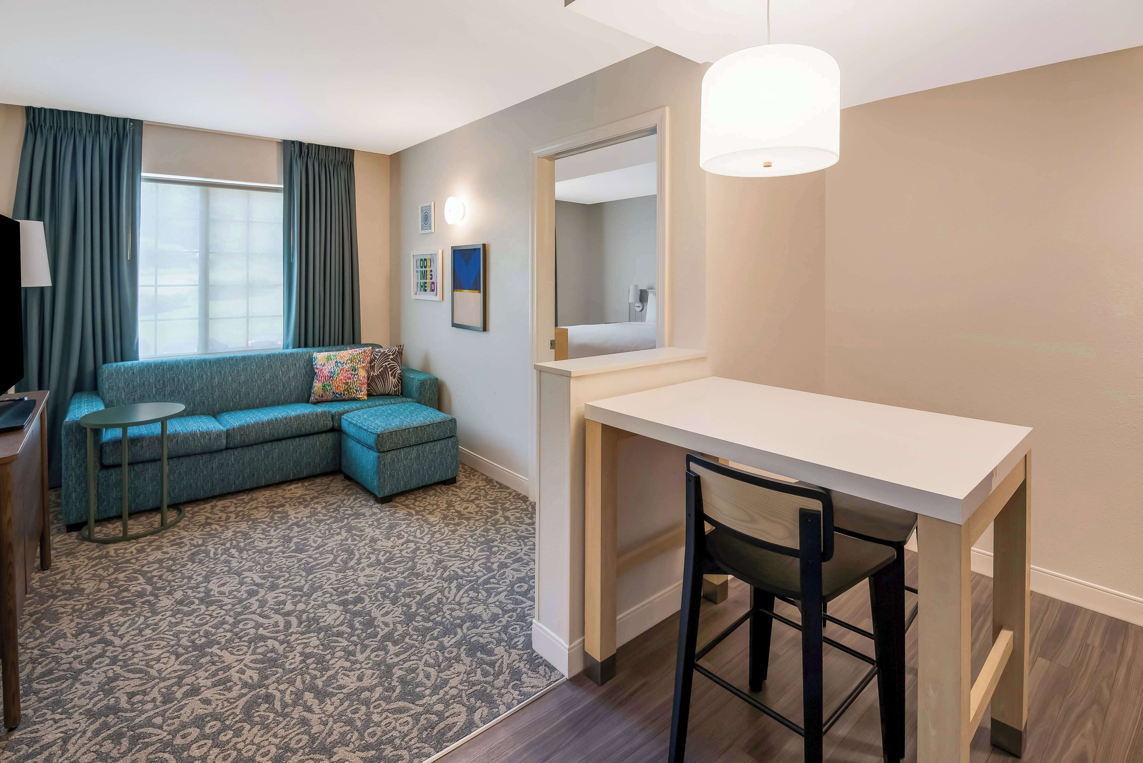 A compact living space with a sofa, a small round table, a desk with a chair, a mirror, and a hanging light fixture.