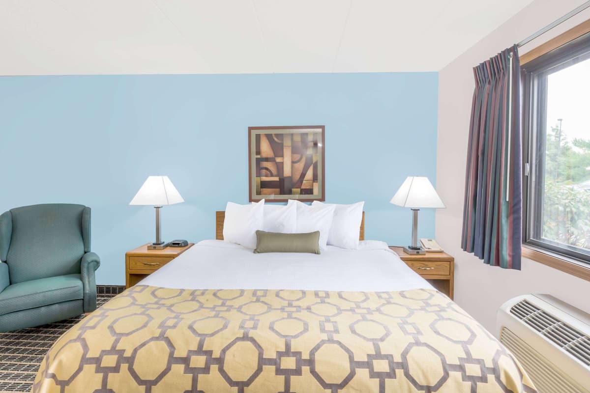 Habitación de hotel con cama, dos lámparas de noche, una silla, una obra de arte enmarcada sobre la cama y una ventana con cortinas.