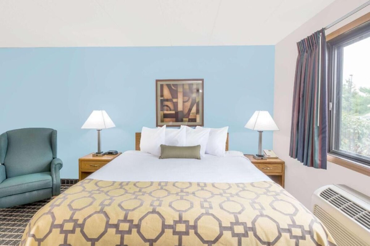 Hotelkamer met een bed, twee bedlampen, een stoel, een raam met gordijnen en een schilderij aan de muur.