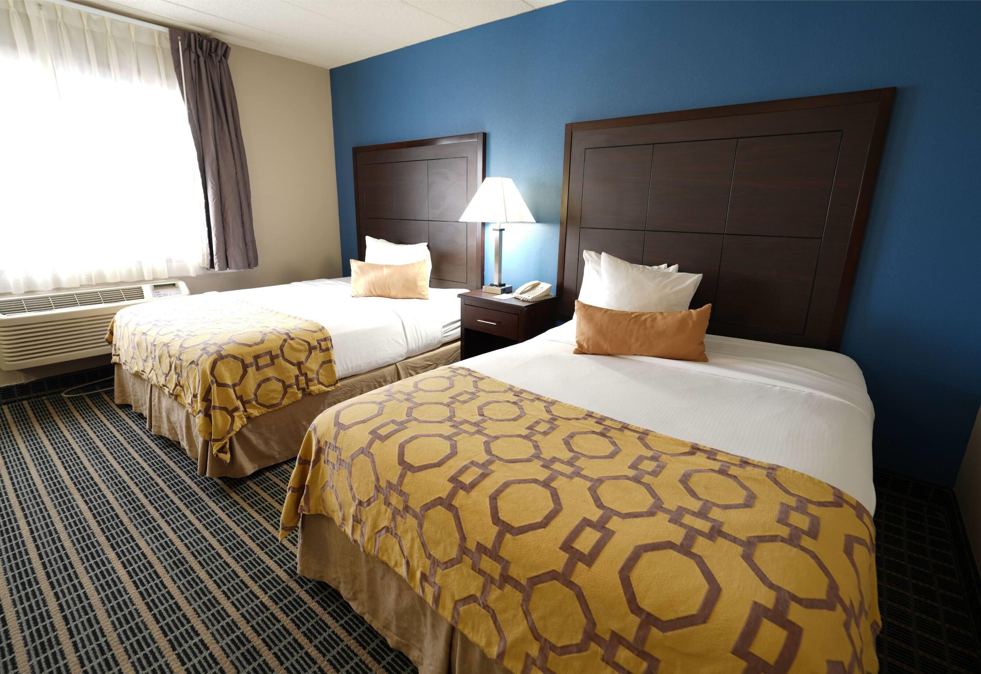 A hotel room with two beds, a nightstand with a lamp, and a window with curtains.