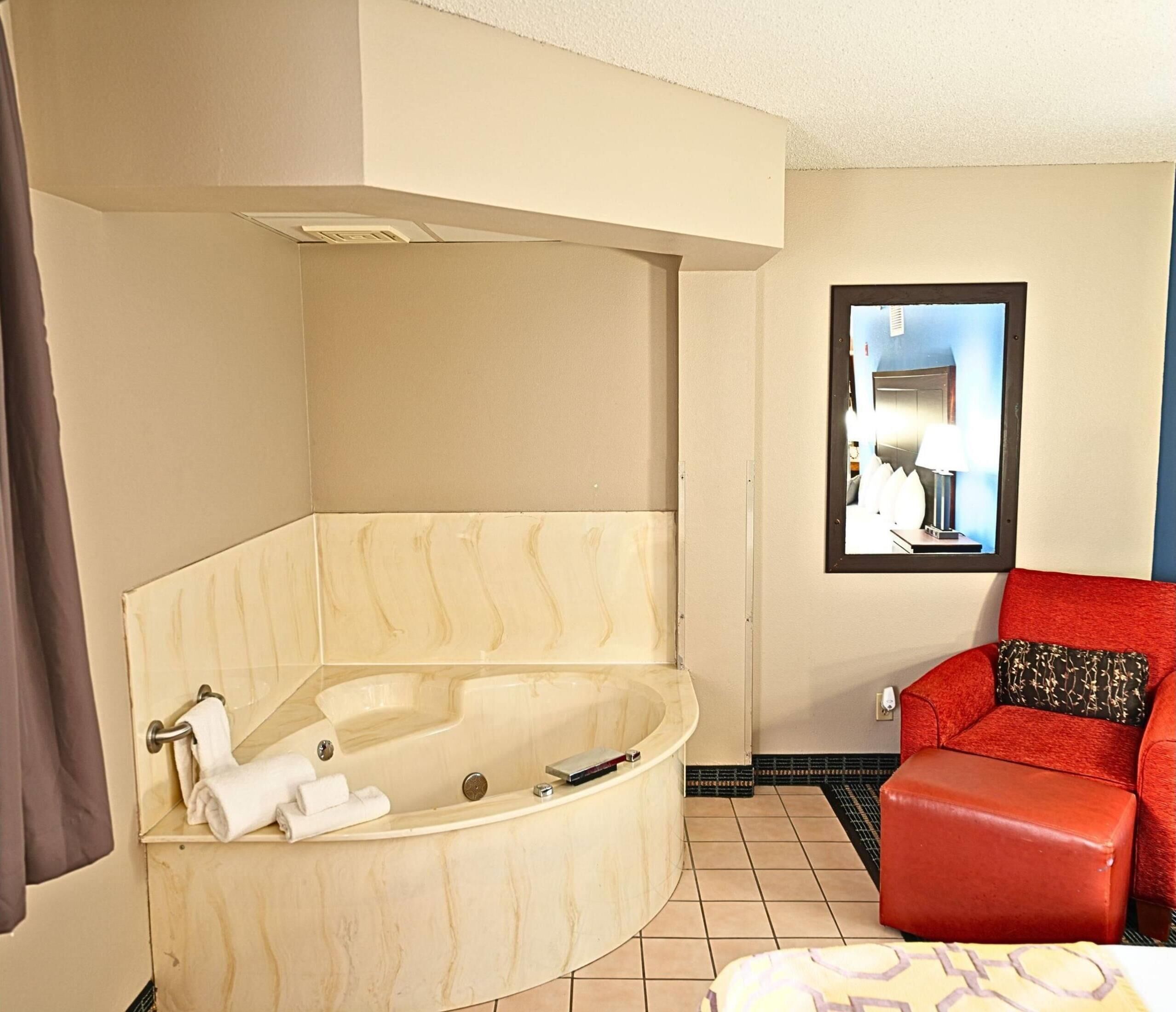 A bathroom with a large bathtub, a red chair, and a mirror reflecting a room with a bed and a nightstand.