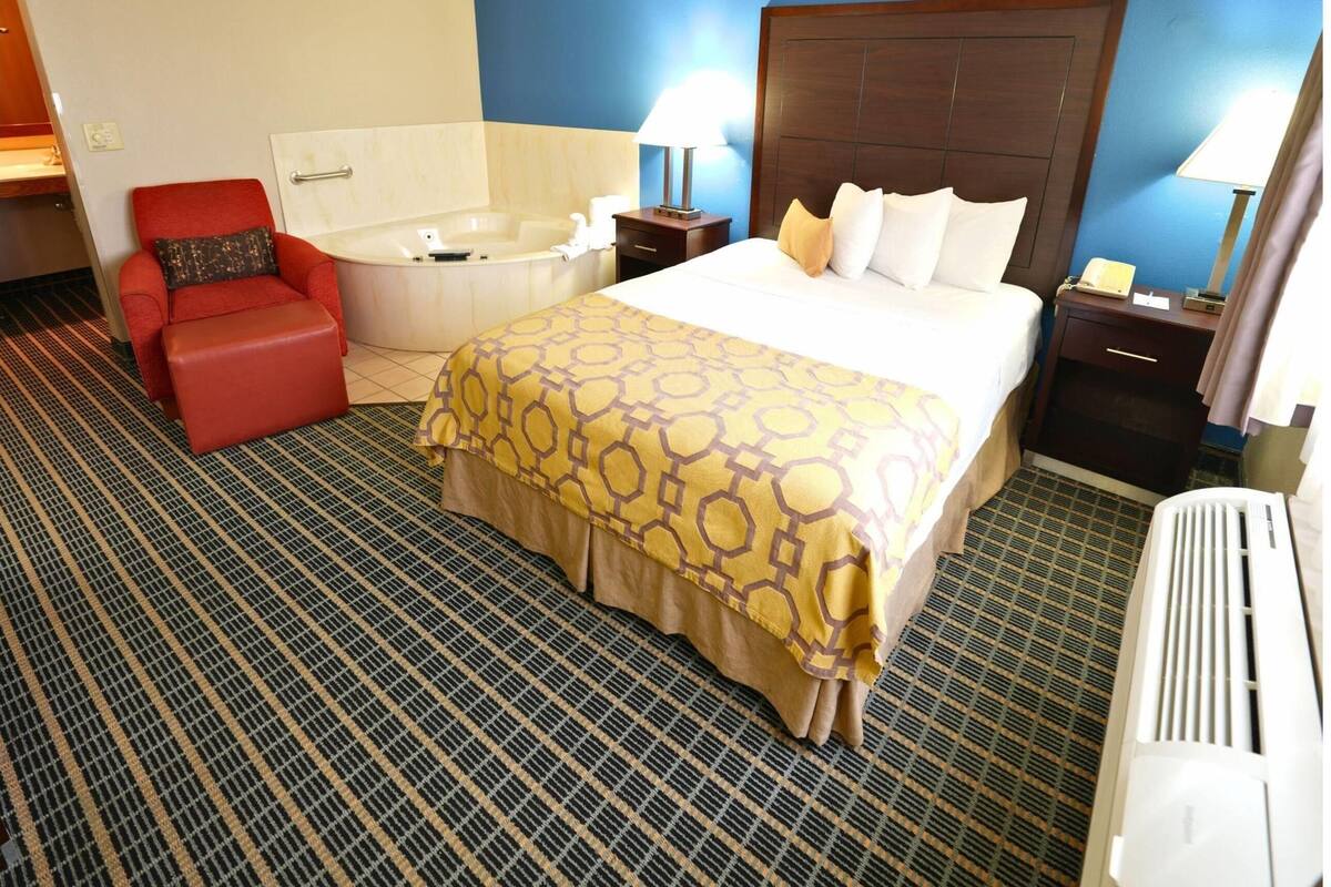 Habitación de hotel con una cama grande, un sillón rojo, una bañera y una pequeña mesa con lámpara.