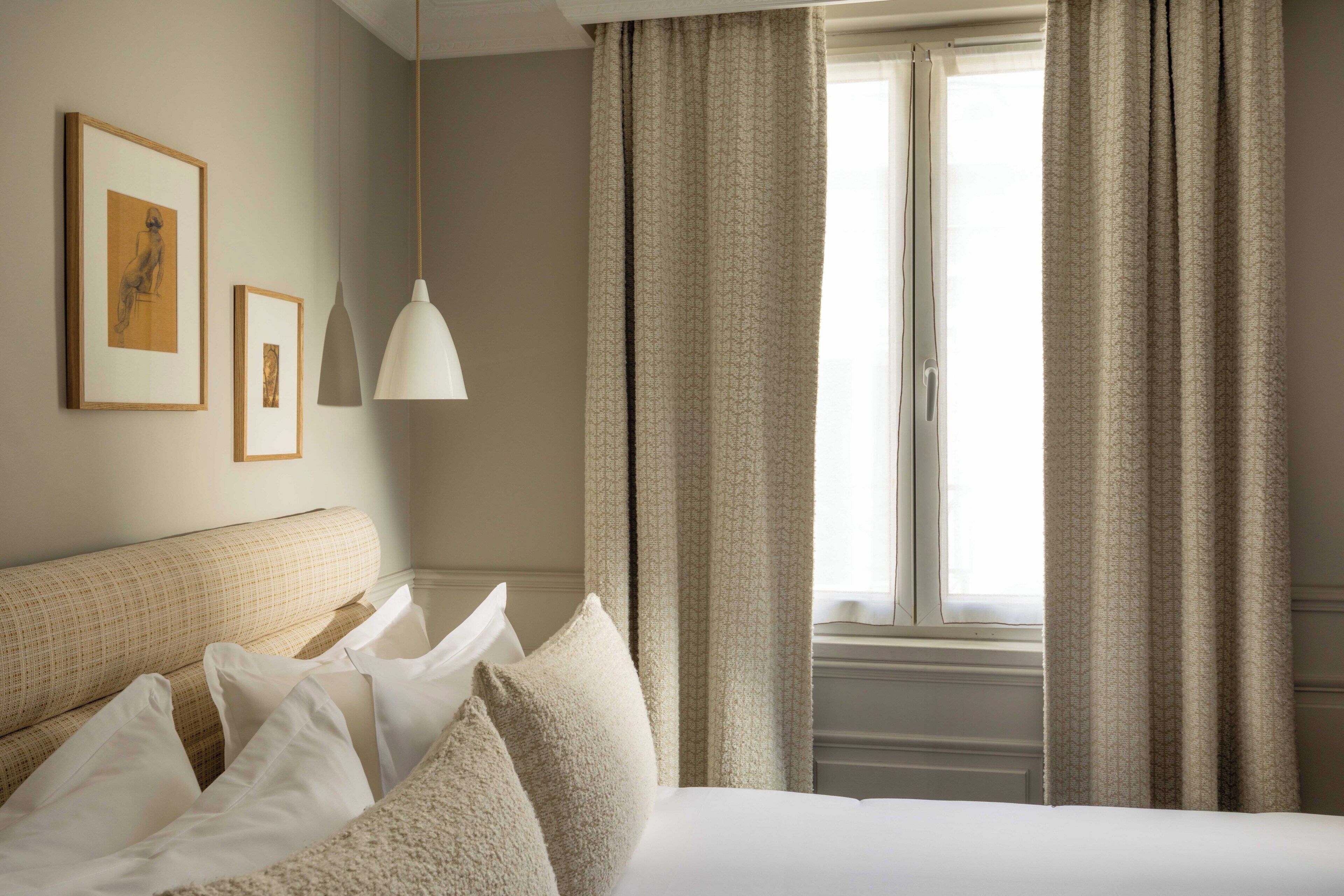 Quarto de hotel com cama, almofadas, luminária pendente e cortinas.