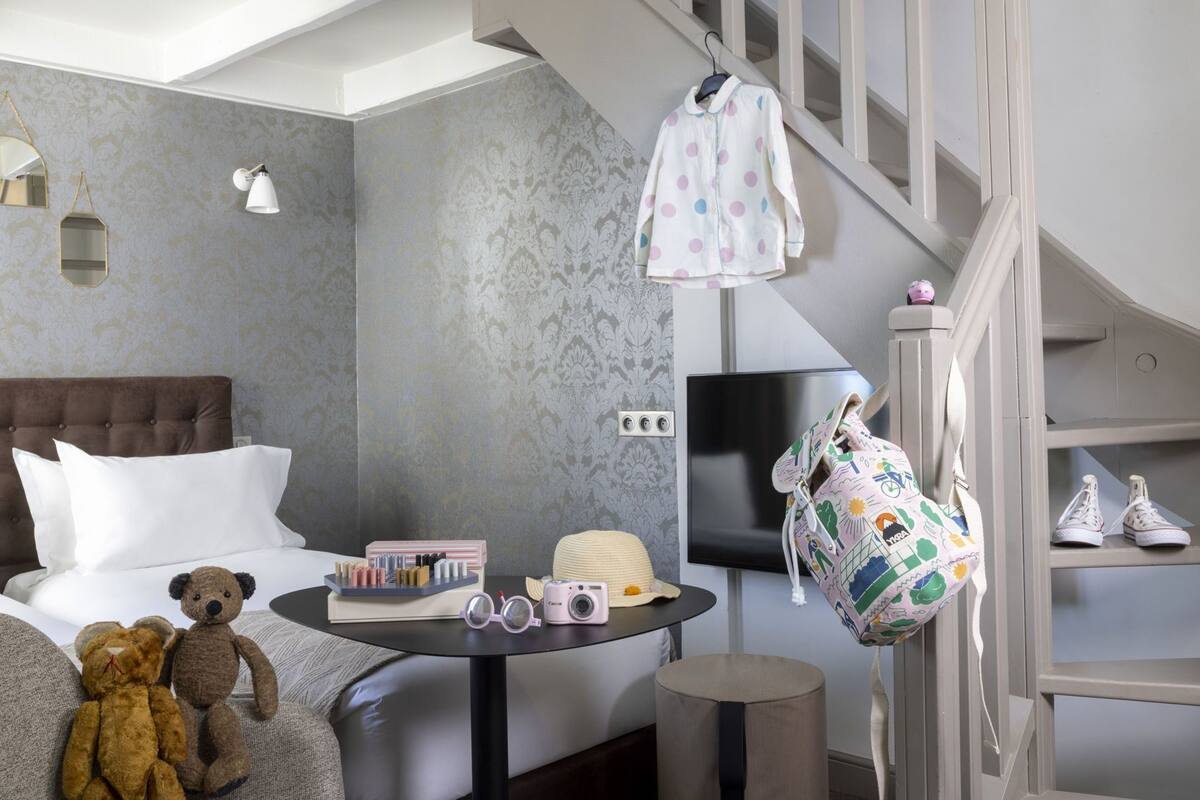 Un dormitorio con una cama, una mesa redonda con objetos, un oso de peluche, una escalera y un televisor.