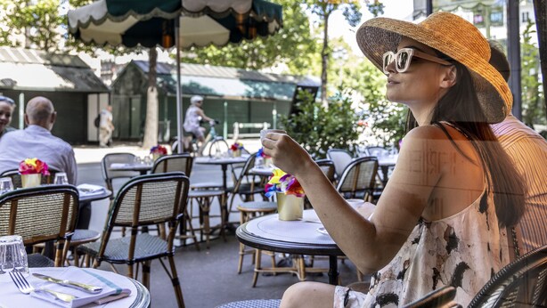 An outdoor cafe with wicker chairs and tables, a green umbrella, and a person sitting at a table. - Hôtel Madeleine Haussmann (Paris)