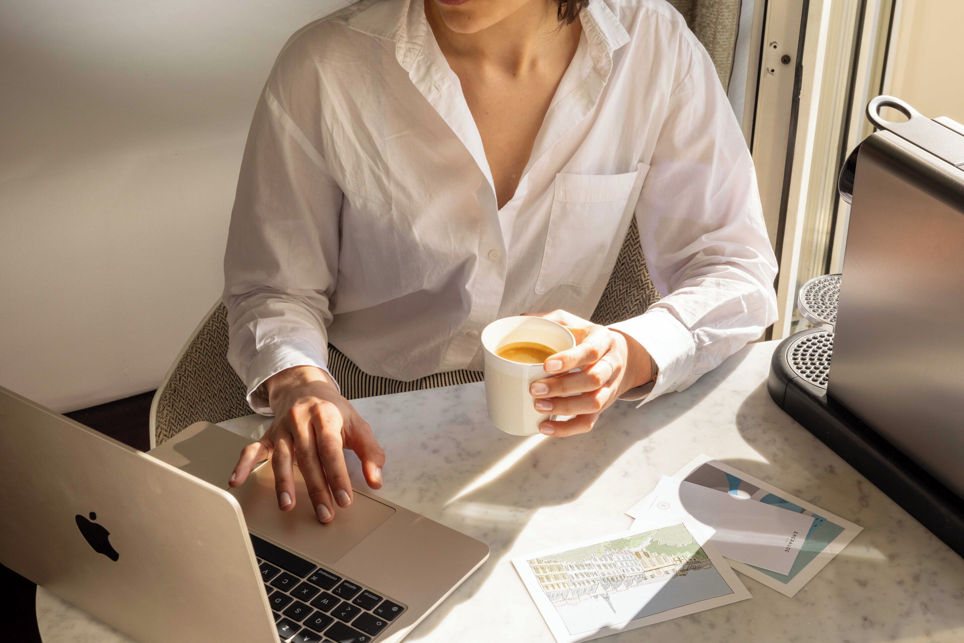 A person using a laptop at a table with a cup of coffee, papers, and a speaker.