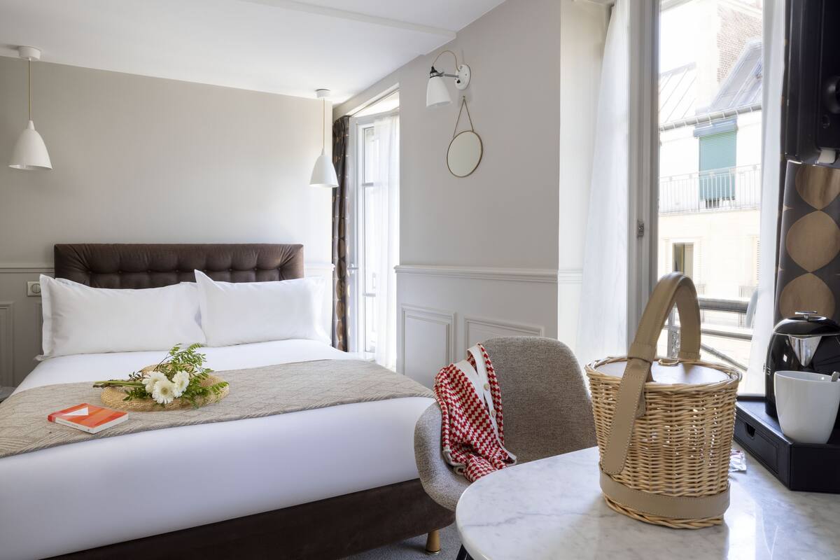 Habitación de hotel con cama, una silla, una mesa, una cesta de mimbre y ventana con vista a edificios.