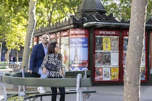 Ein Paar sitzt auf einer Bank in einem Park, im Hintergrund ist eine Bushaltestelle und Werbung zu sehen.