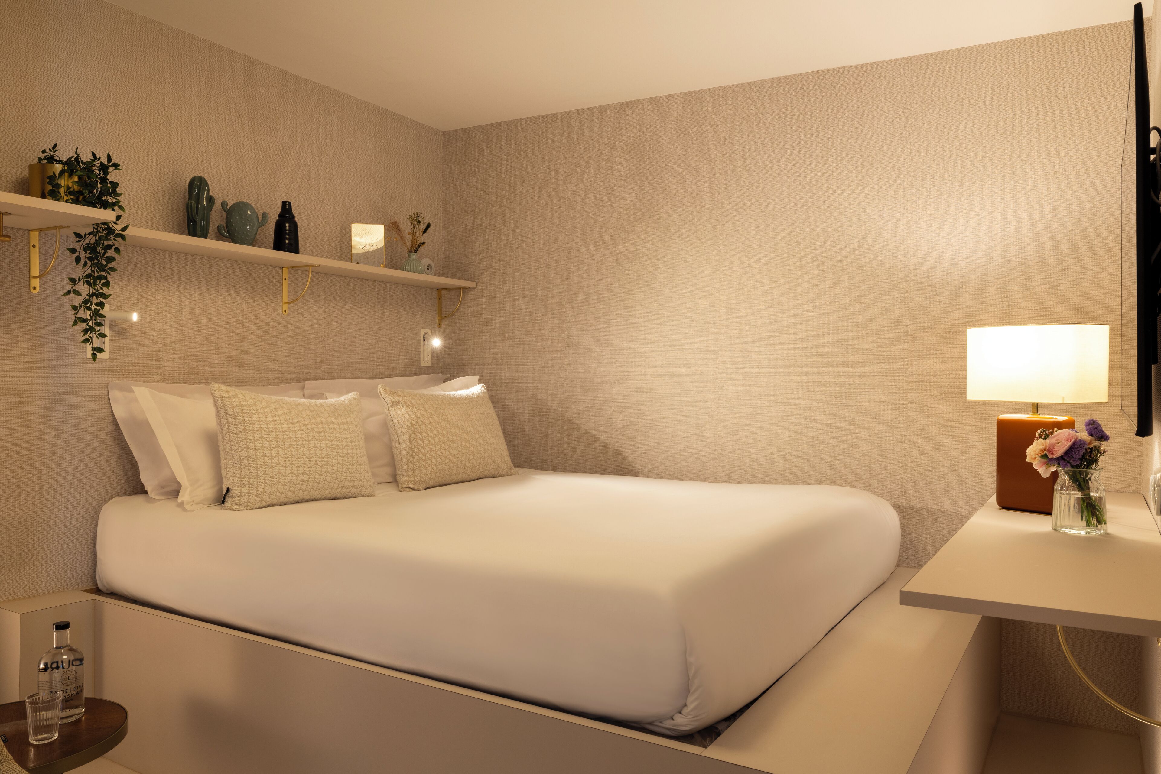 Un cuarto de hotel ordenado, con una cama, mesitas de noche, una lámpara y objetos decorativos en estantes.