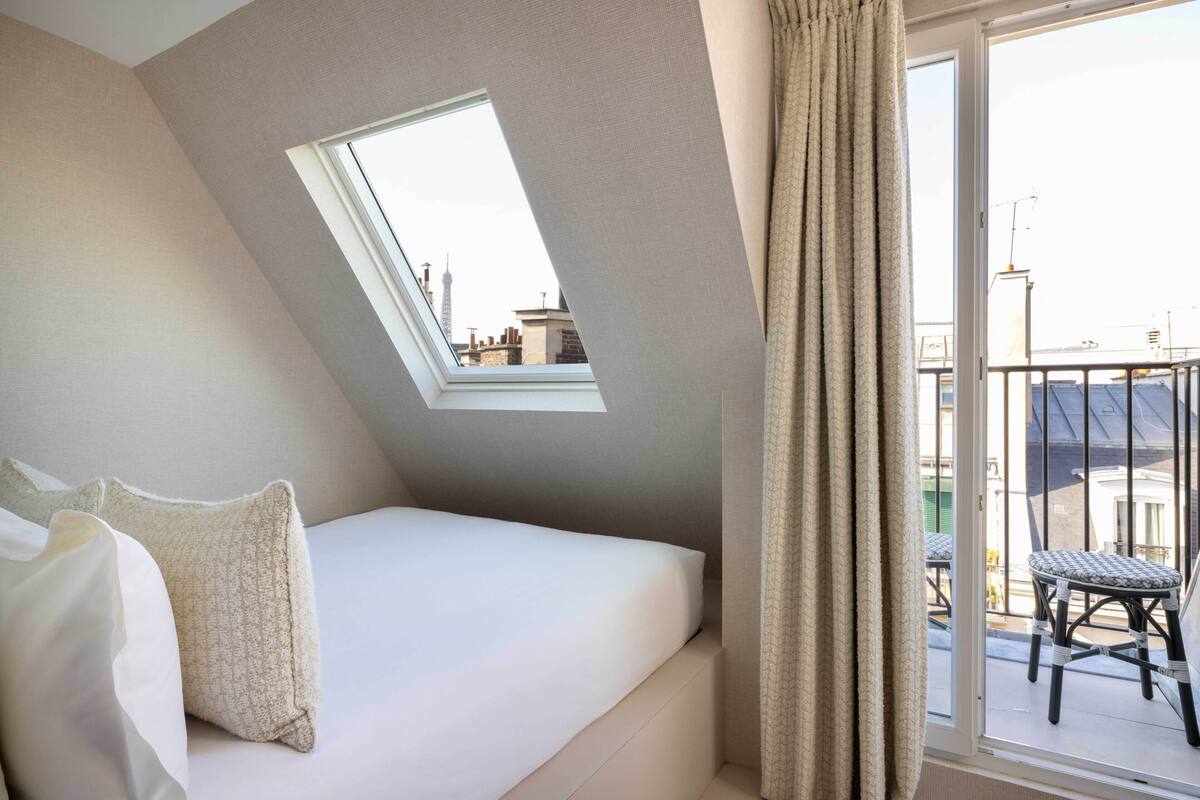 Un dormitorio con techo inclinado, tragaluz, cama con ropa de cama blanca y vistas de la ciudad desde un balcón.