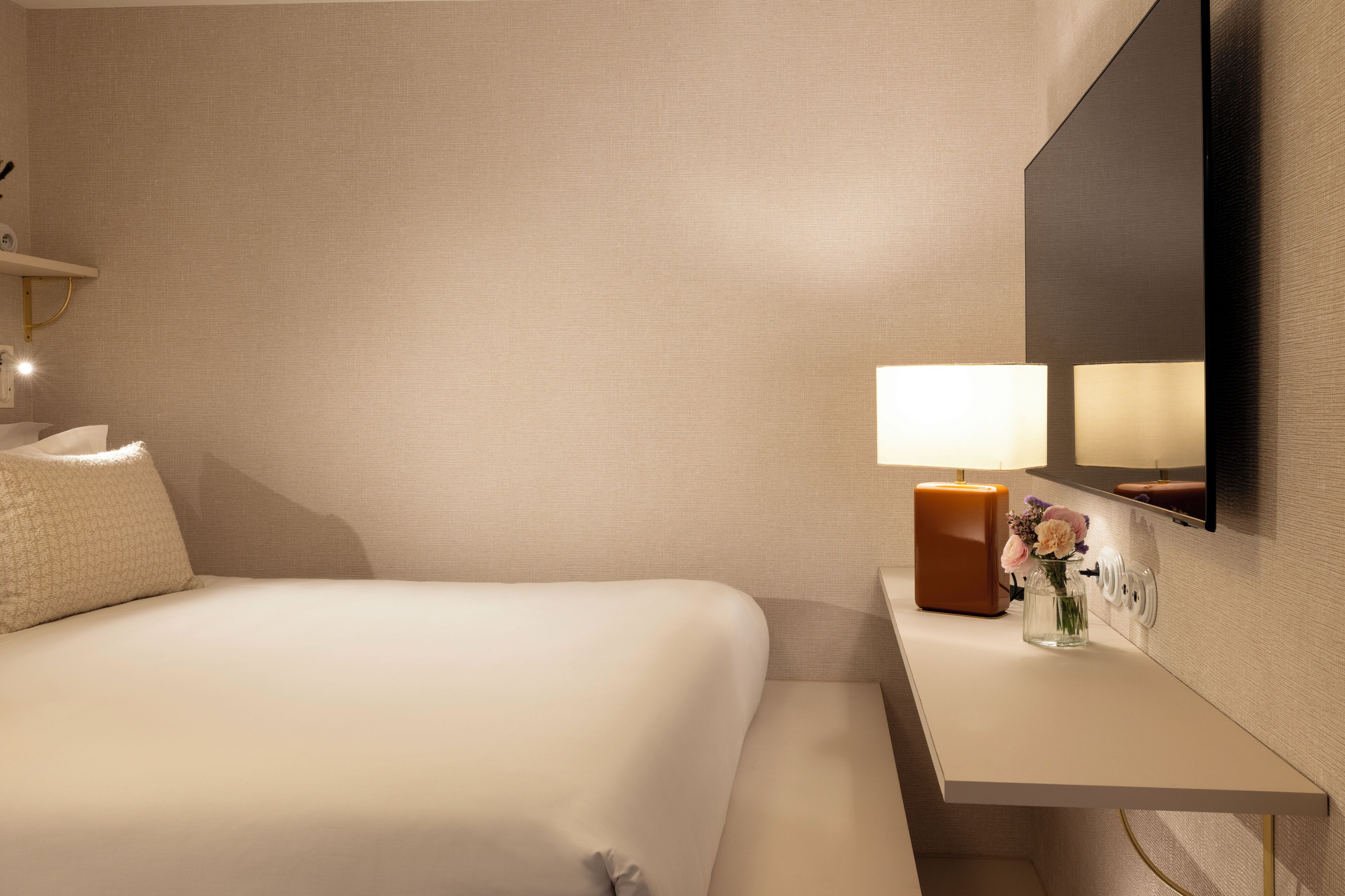A hotel room with a bed, bedside table, lamp, vase with flowers, and a mirror.