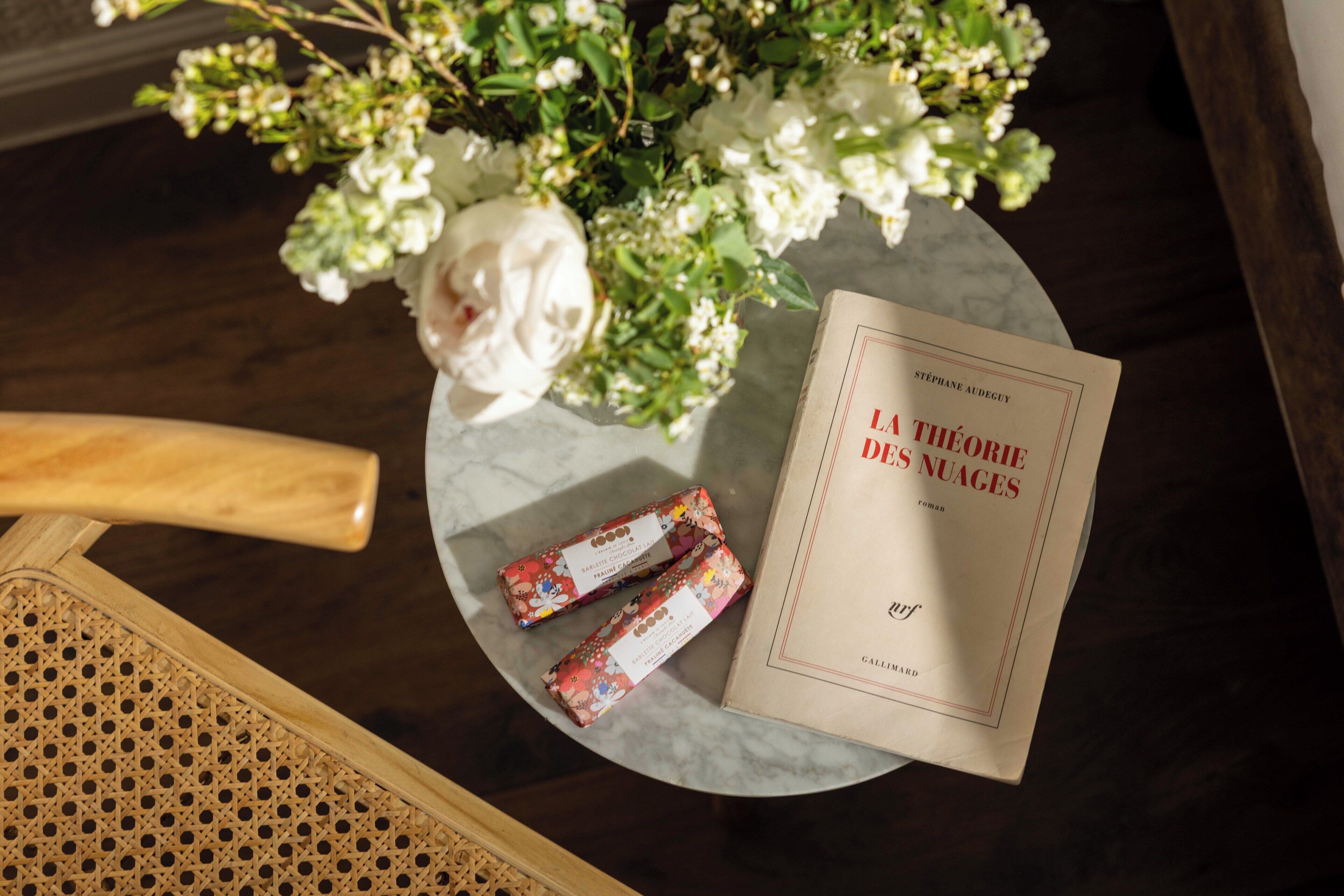 A book titled 'La Théorie des Nuages' by Stéphane Audeguy on a table with a white flower arrangement and two chocolate bars.