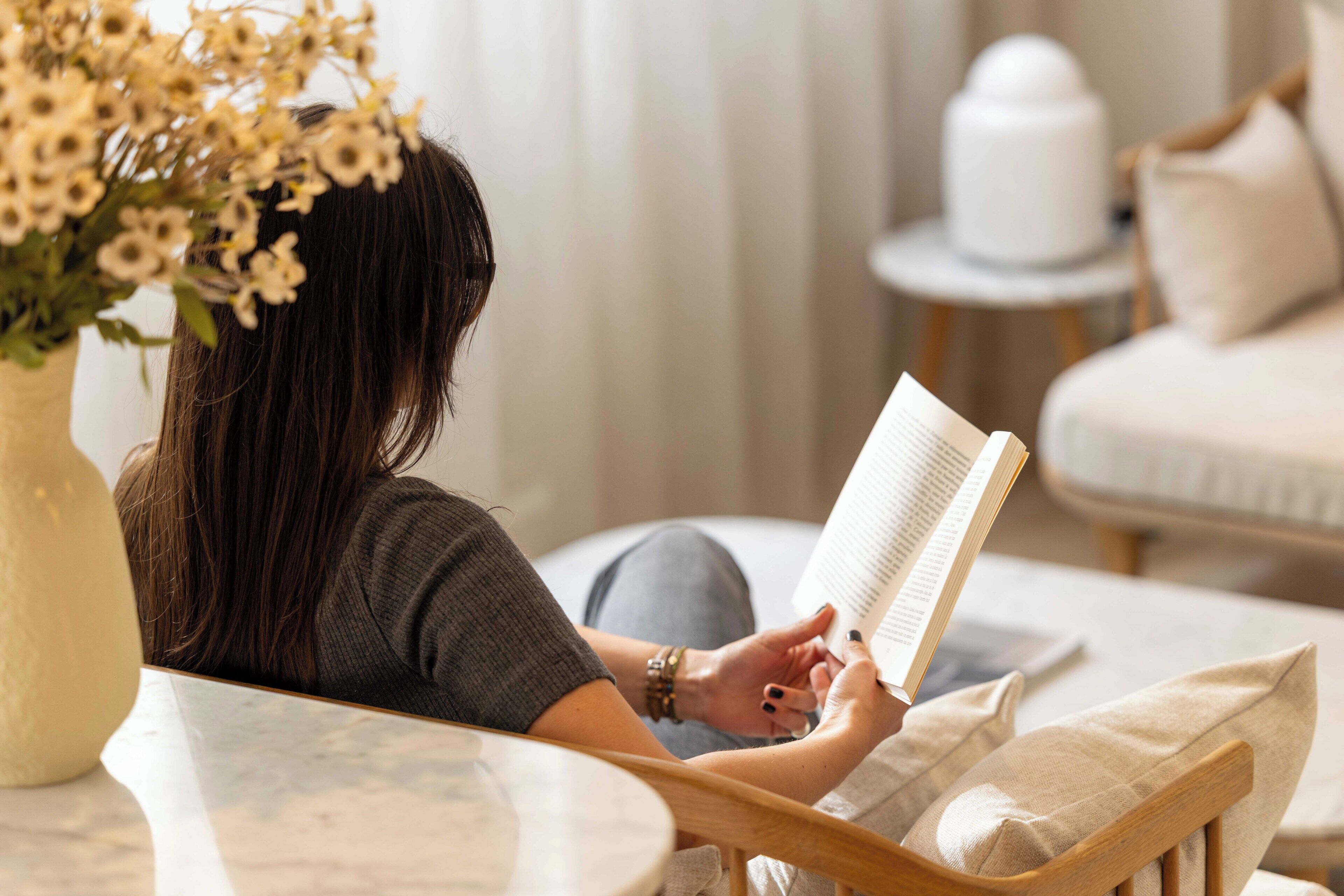 A person reading a book in a room with a round table, a chair, and a vase with flowers.