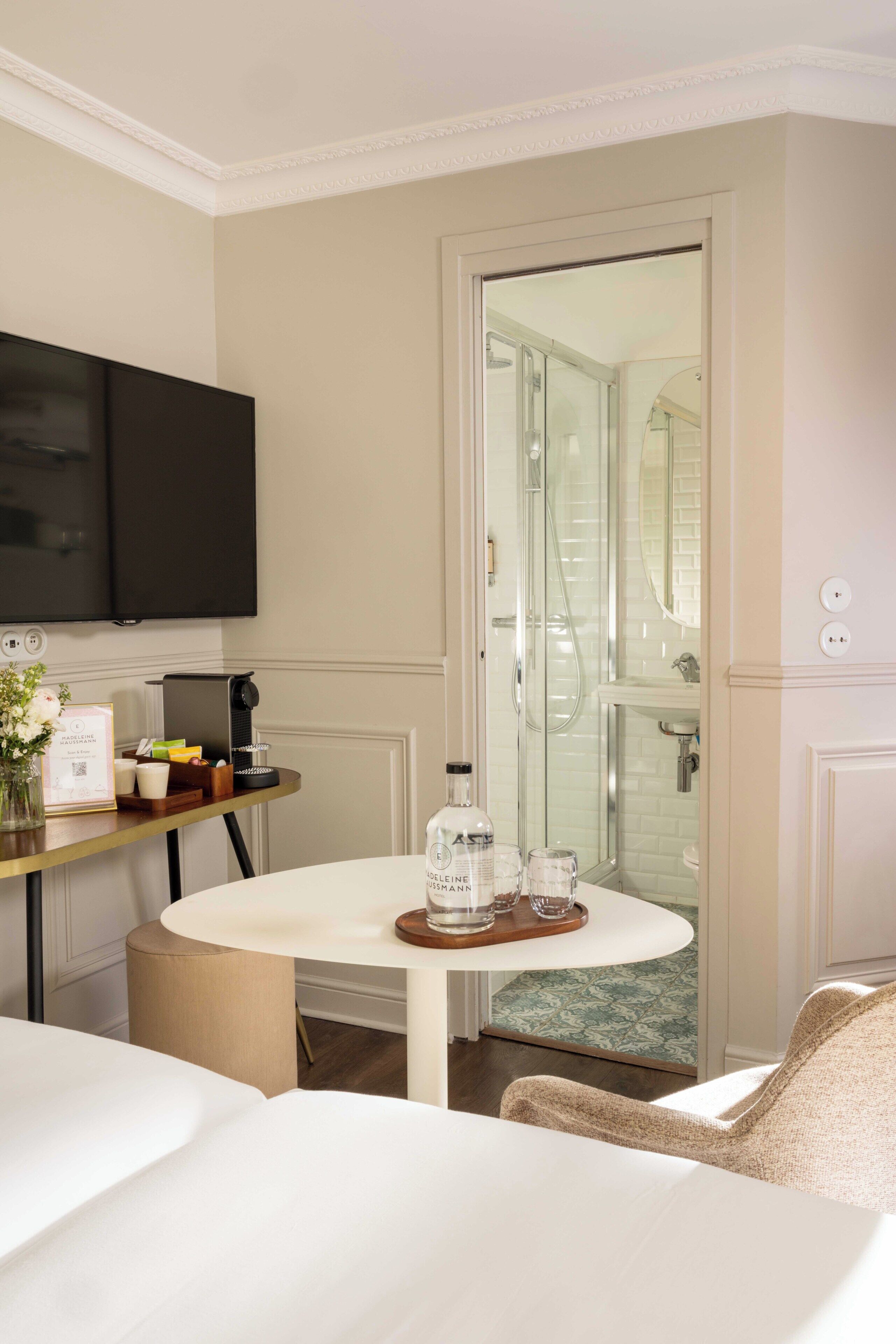 A hotel room with a bed, a round table with a tray, a TV, a shower area, and a small desk with a coffee maker.
