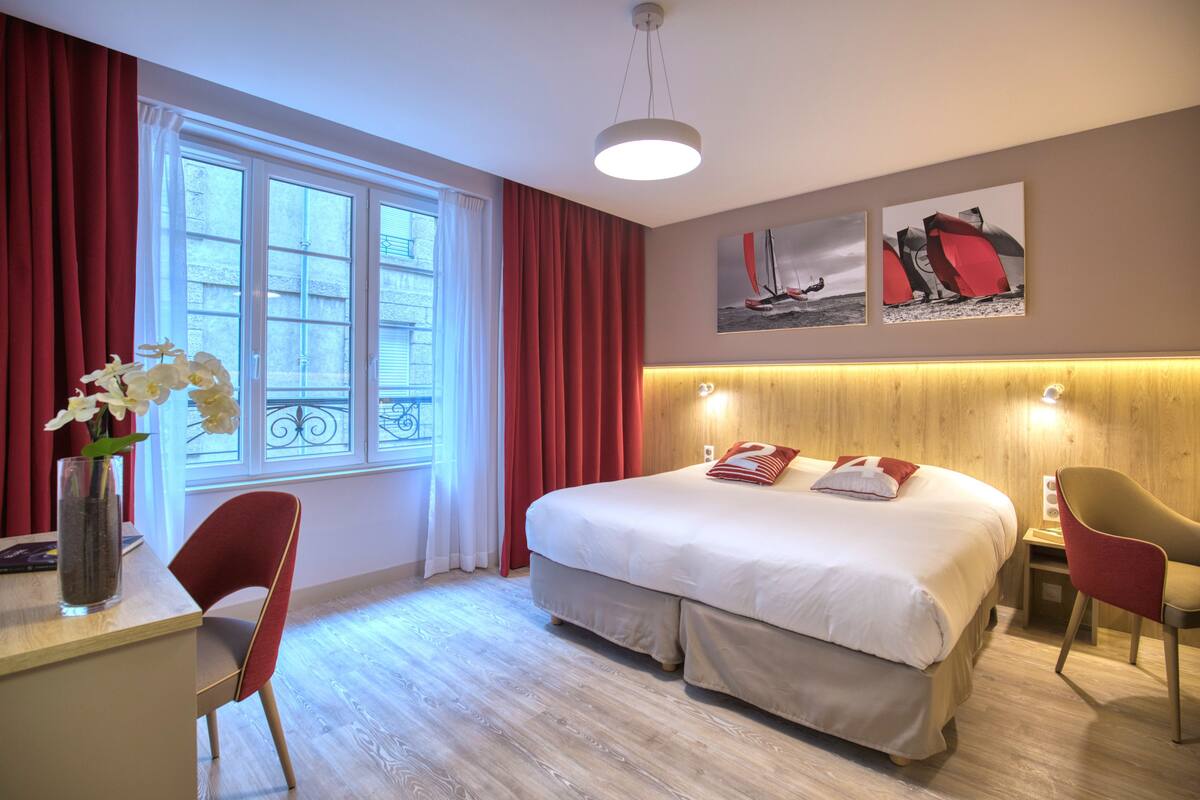 Une chambre d’hôtel avec un grand lit, un bureau, une chaise, un vase avec des fleurs et une fenêtre avec des rideaux.