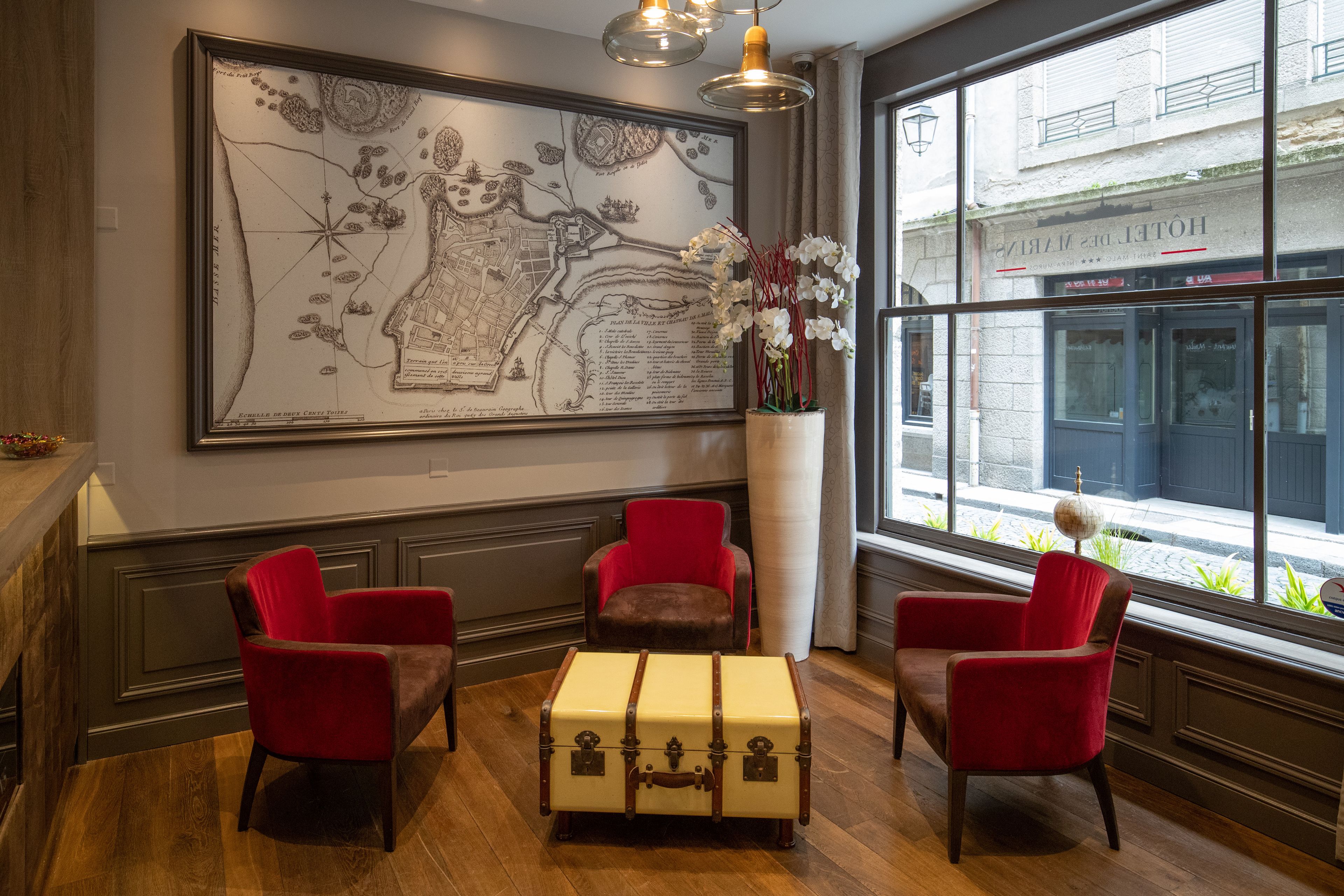 Une pièce avec une grande carte accrochée au mur, des fauteuils rouges, une vieille valise et une grande fenêtre donnant sur la rue.