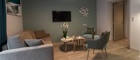 A modern living room with a grey sofa, a small table with a plant, and a grey armchair. A flat-screen TV is mounted on the wall, and a door is visible in the background.