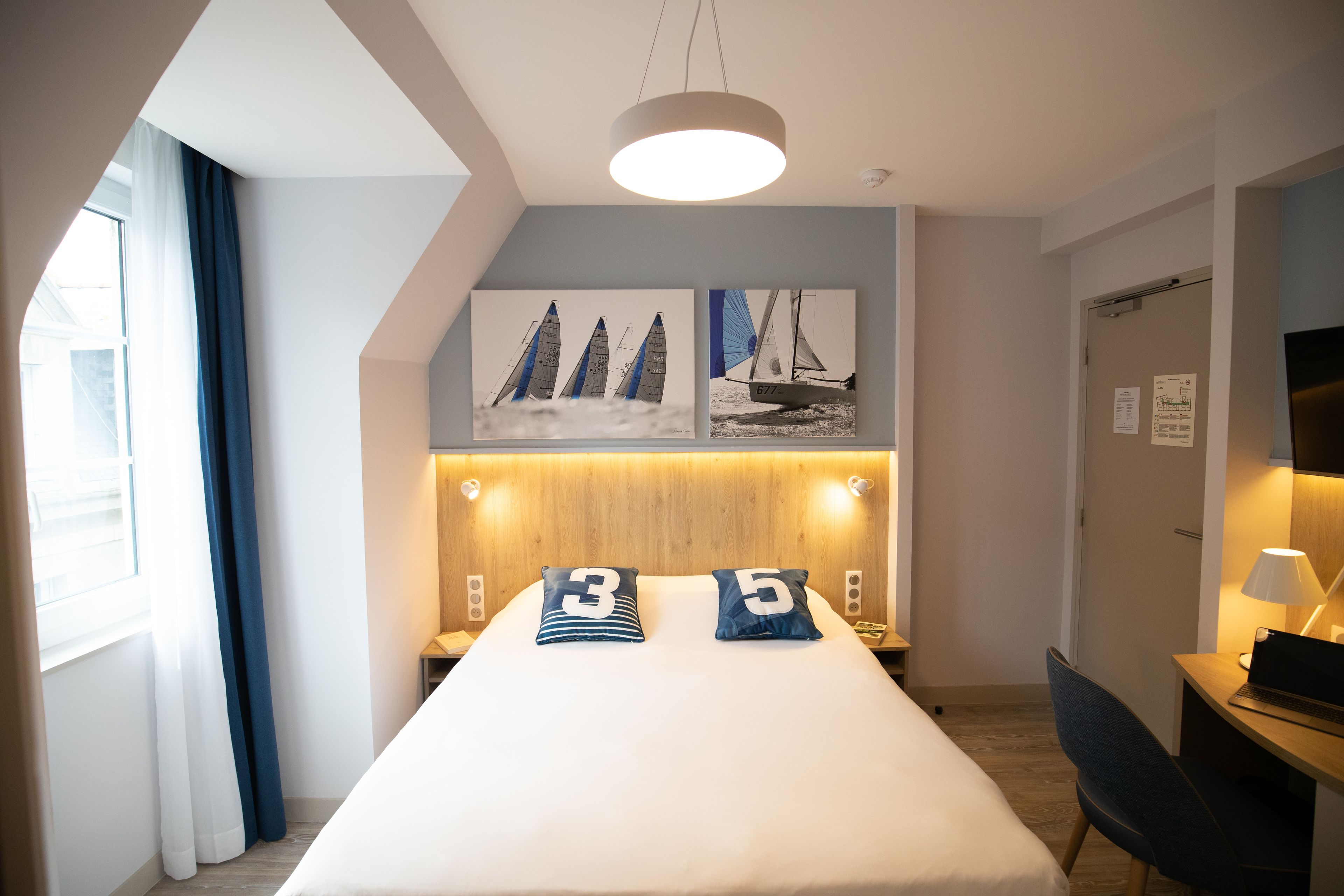 Habitación de hotel moderna con una cama grande, dos almohadas, un escritorio con una computadora portátil, una silla, una lámpara de escritorio y dos cuadros colgados en la pared con barcos a vela.