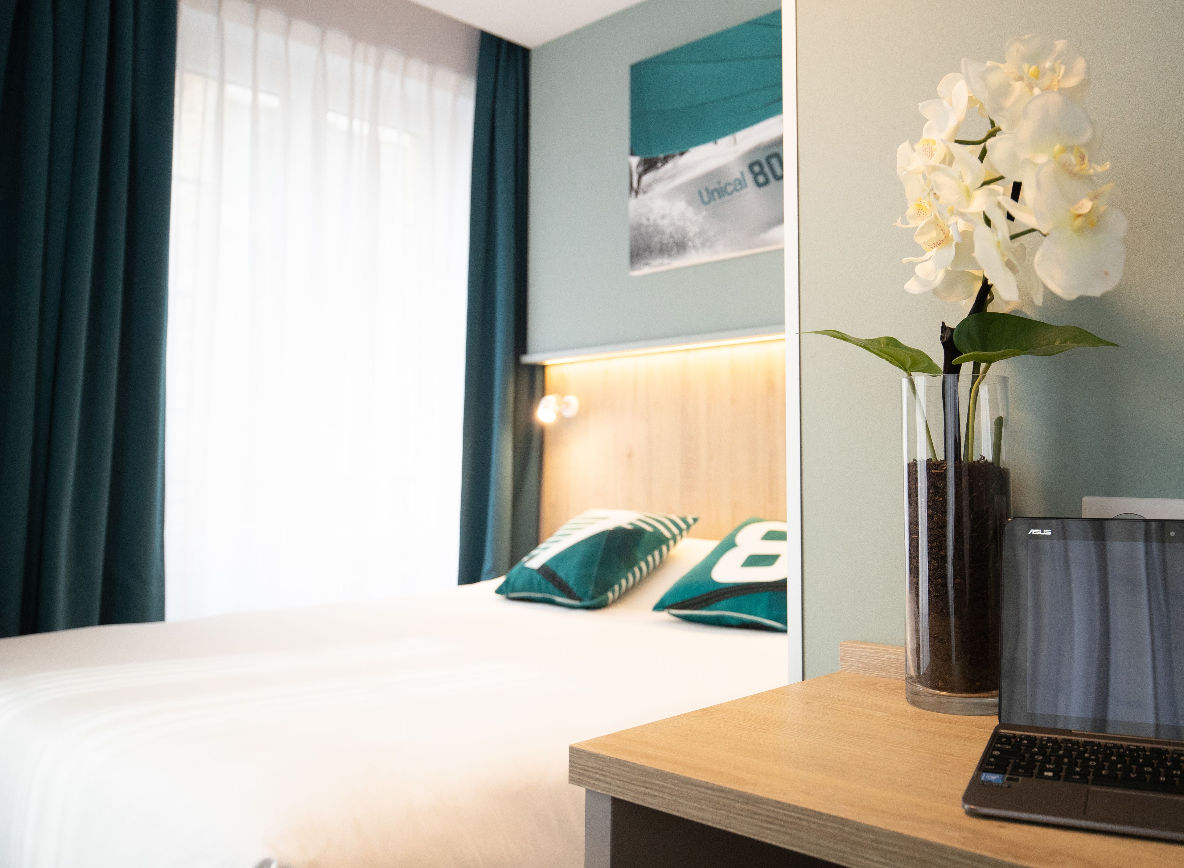 A hotel room with a bed, a laptop on a desk, and a vase with white flowers.