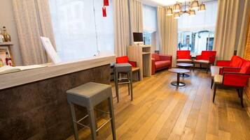 Een moderne hotellobby met een houten vloer, rode en grijze zitplaatsen, een receptiebalie en grote ramen met gordijnen.