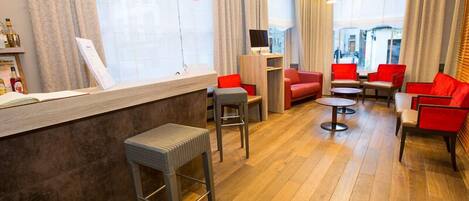 Un hall d’hôtel moderne avec un plancher en bois, des sièges rouges et gris, un comptoir d’accueil et de grandes fenêtres avec des rideaux.