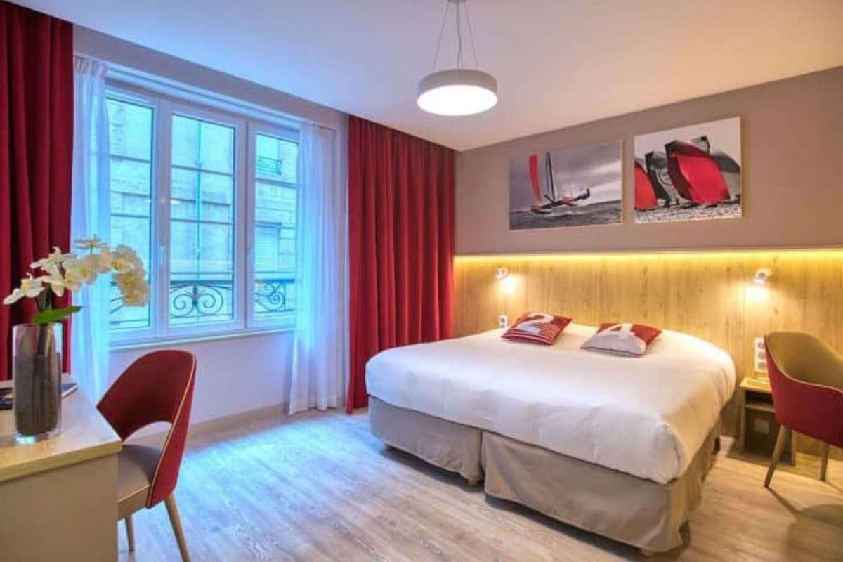 Et hotelværelse med en stor seng, et skrivebord med stol, et lille bord og et vindue med røde gardiner.