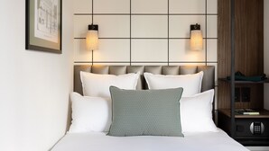 A neatly made bed with white and patterned pillows, a framed architectural drawing on the wall, and a nightstand with a lamp and a small electronic device. - Hotel Moderniste (Paris)