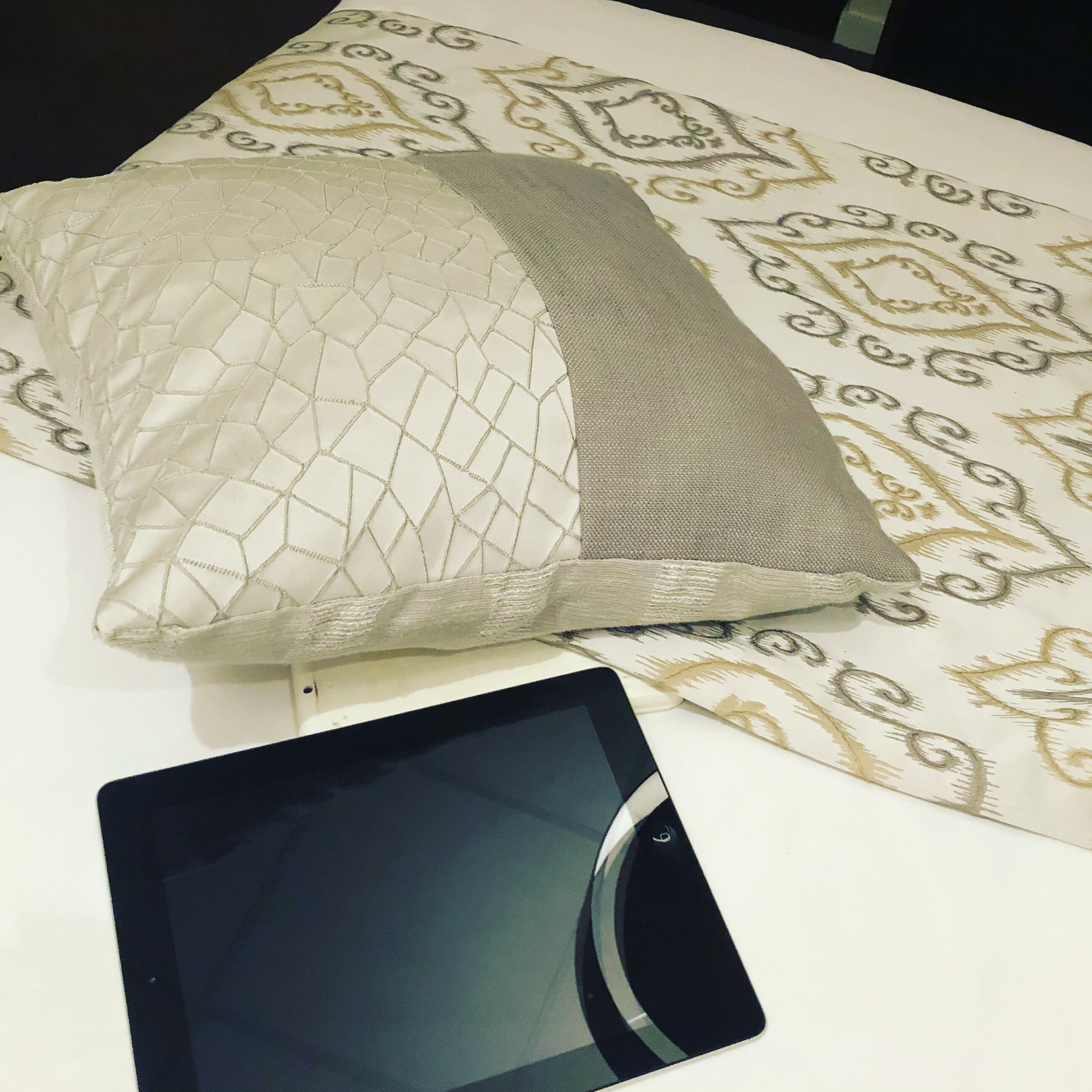 A bed with a patterned bedspread and a pillow with a quilted design. A tablet is placed on the bed.