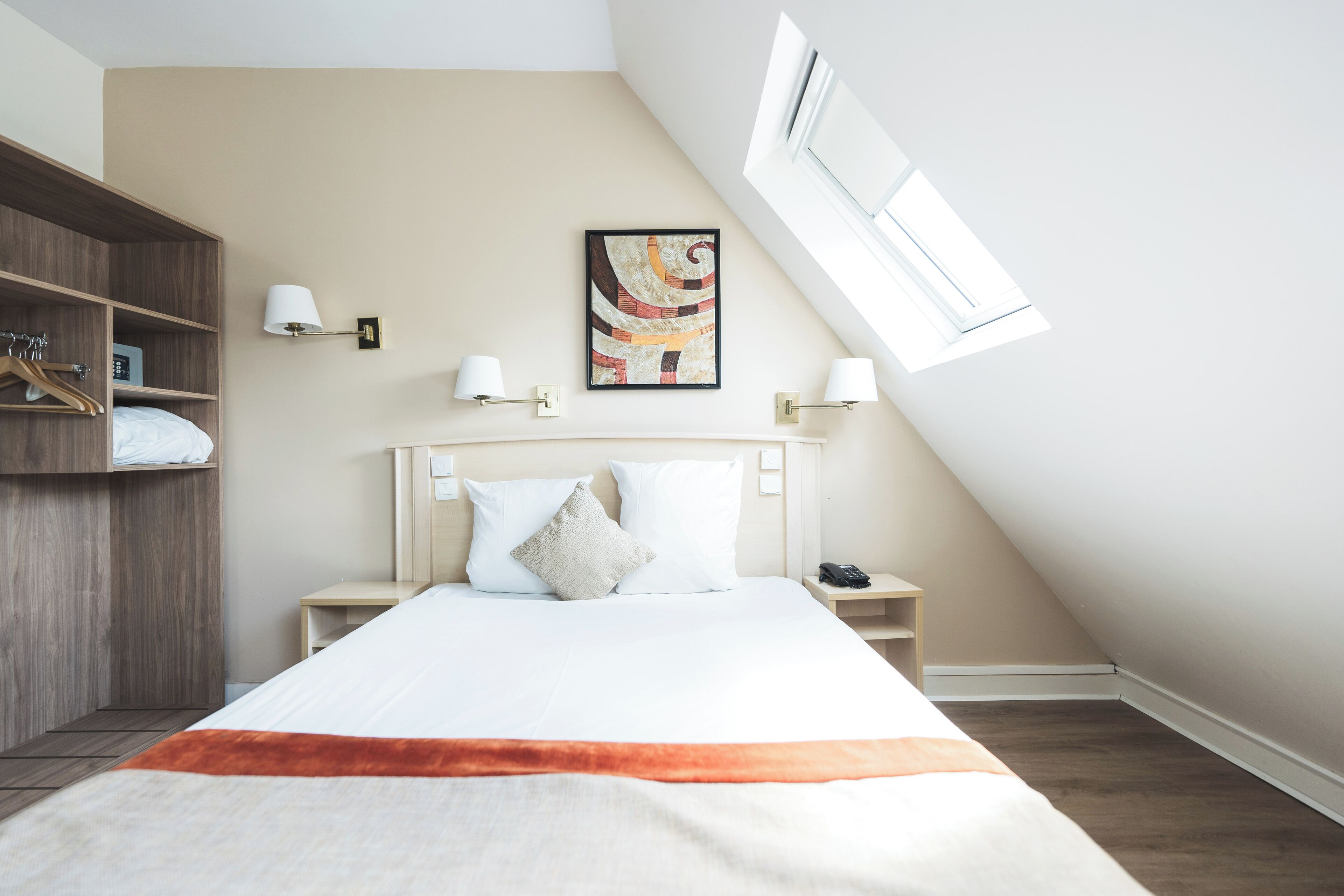 A modern hotel room with a large bed, a wardrobe, a bedside table, and a painting on the wall.