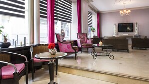 En moderne hotellobby med et siddeområde, store vinduer med persienner, en lysekrone og et fladskærms-TV på væggen.