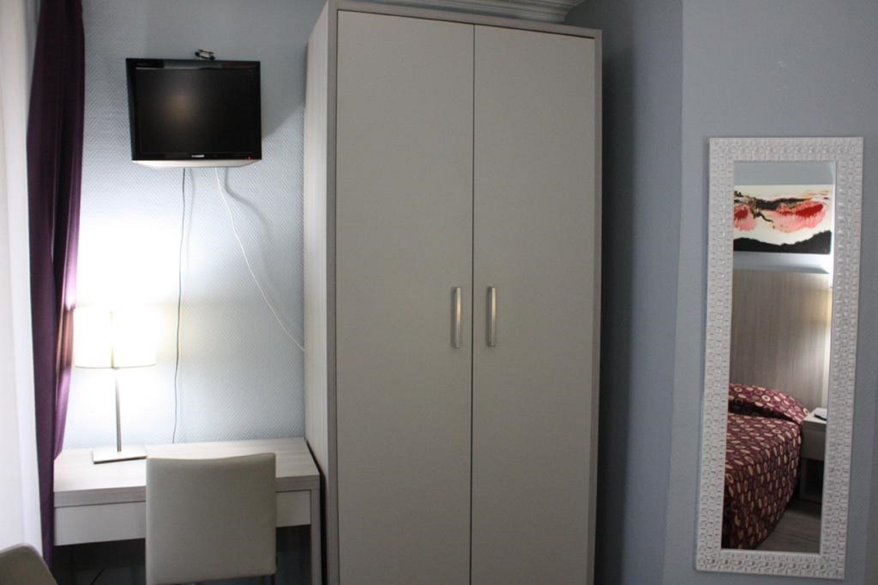 A hotel room with a wardrobe, a desk, a chair, a television mounted on the wall, and a mirror with a decorative frame.