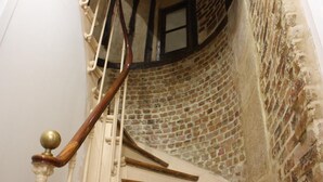 Escada em espiral com degraus de madeira e corrimão de latão, que leva a um quarto com paredes de tijolos aparentes e uma janela.