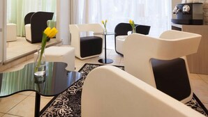 Quarto de hotel moderno com área de estar, incluindo cadeiras brancas e pretas, uma mesa de centro de vidro e um vaso com flores amarelas.