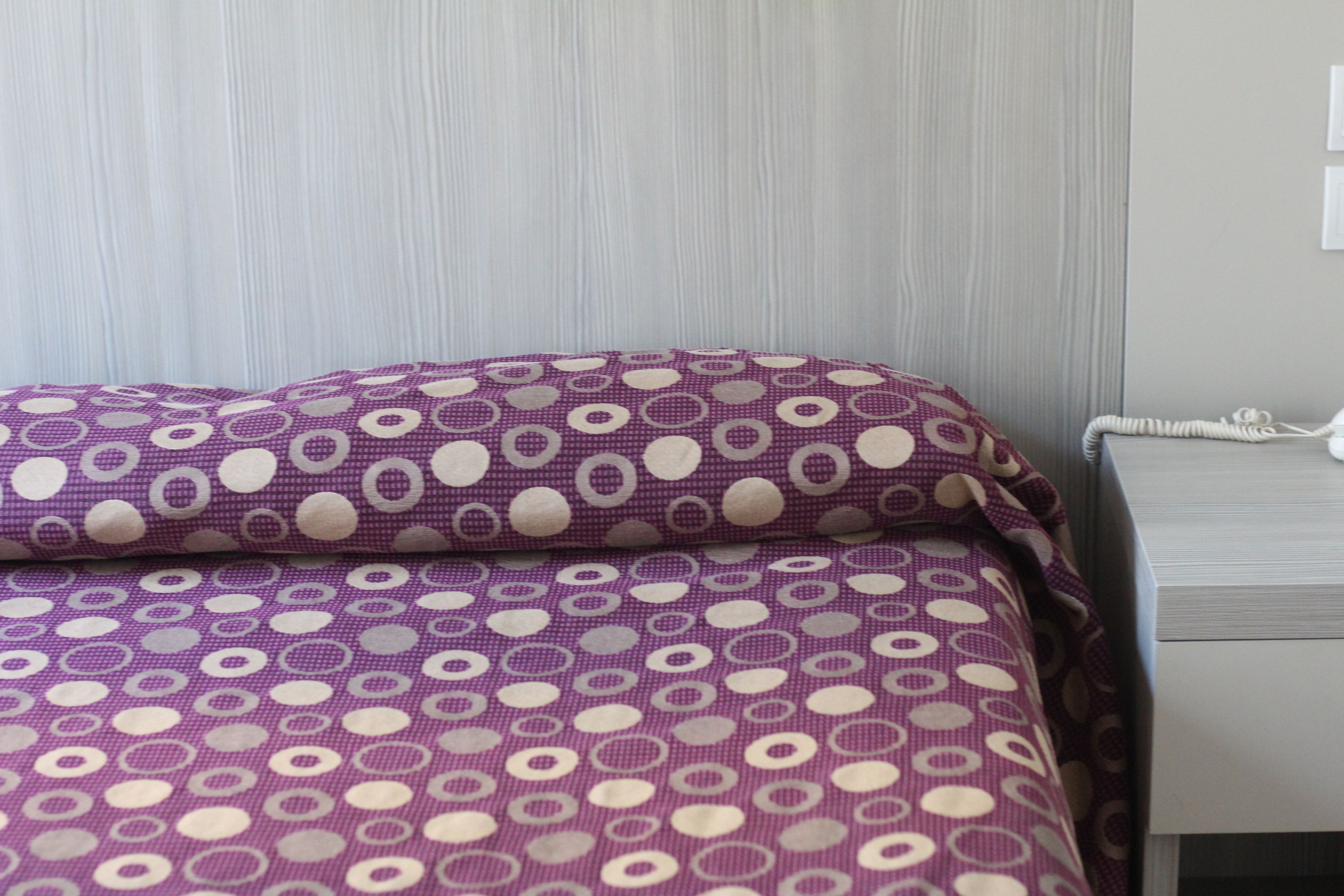 Una cama con una colcha morada y blanca de lunares, una mesita de noche blanca con un teléfono, y una pared de color claro con una ventana.