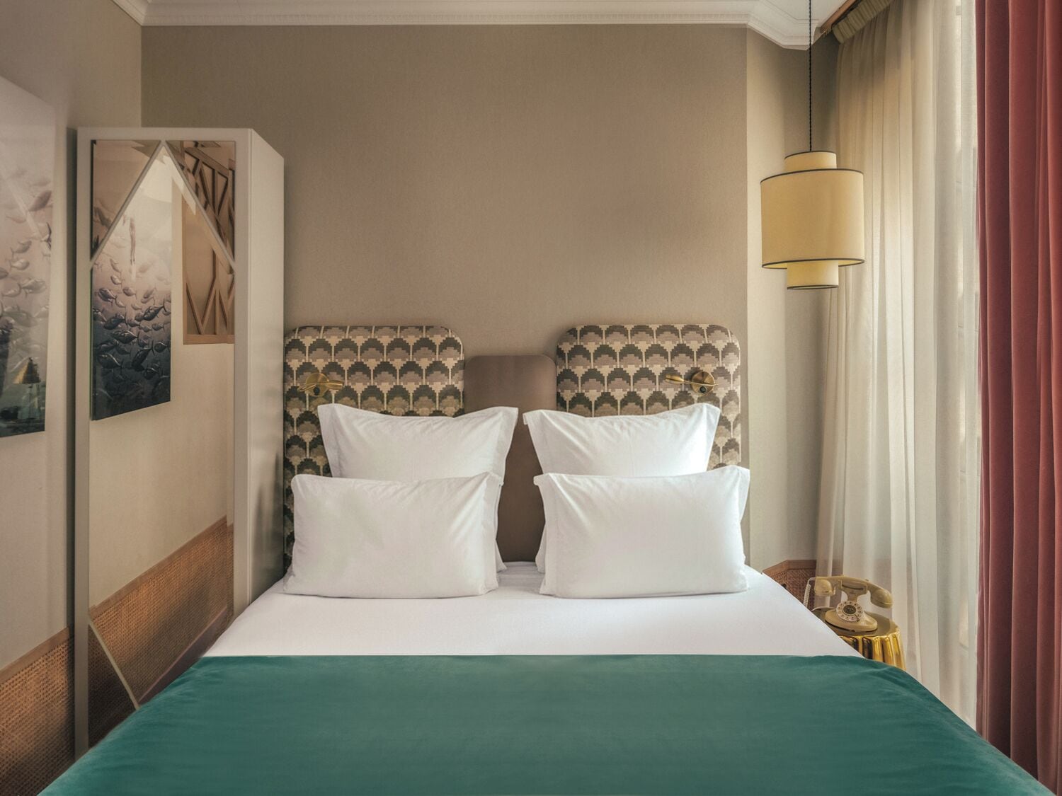 A hotel room with a large bed, two pillows, a green bedspread, a headboard with a patterned design, a wall-mounted artwork, a bedside lamp, and a vintage telephone on a side table.