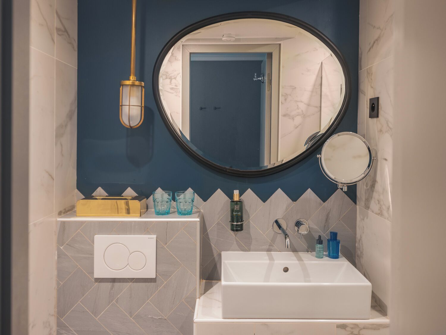 A bathroom with a white sink, a round mirror, and a rectangular mirror above the sink. There is a towel rack with a yellow towel holder and a white switchboard.